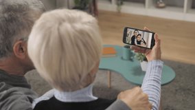 old couple sitting on sofa having a video chat with grandchildren,back view of old person holding smart phone talking with children using videocall technology
