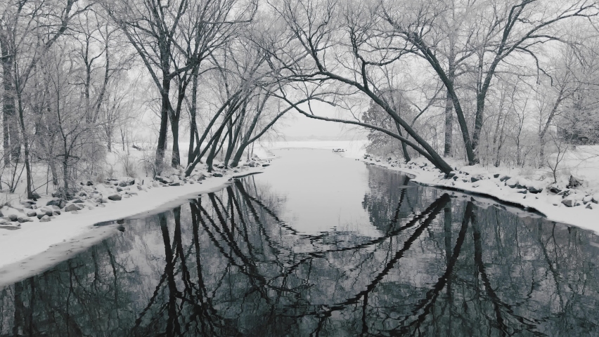 Camera moving low over the water of a winter river and under a canopy of snowy trees emerging into a white barren frozen lake.