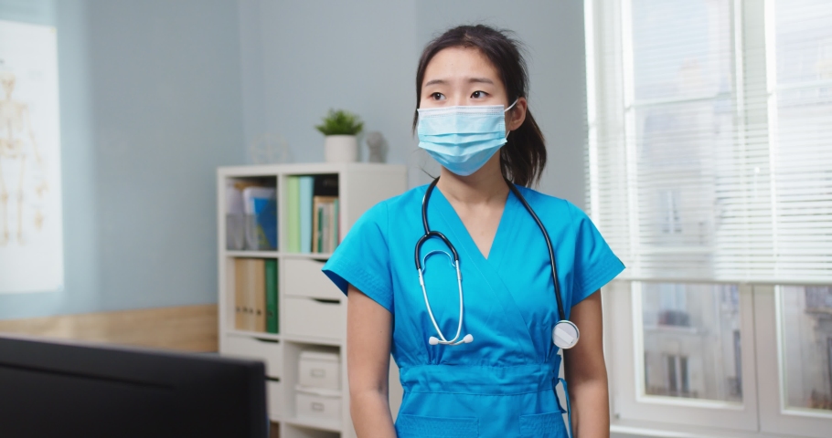 Portrait of good-looking Asian woman in blue uniform in hospital office. Adult female nurse in medical protective mask looking at camera and smiling. Pandemic, Virus, medicine, helath concept.