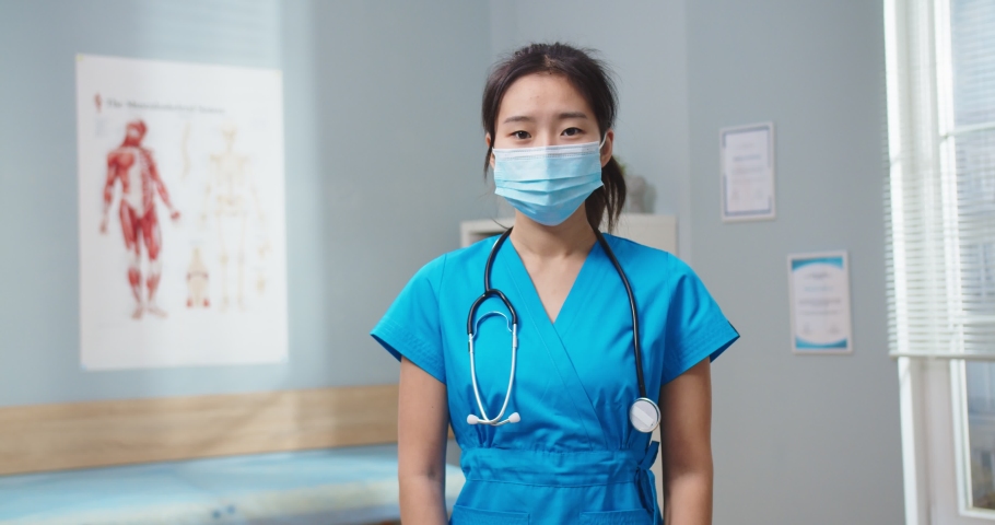 Portrait of good-looking Asian woman in blue uniform in hospital office. Adult female nurse in medical protective mask looking at camera and smiling. Pandemic, Virus, medicine, helath concept.