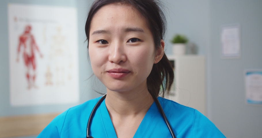 Close up of attractive Asian woman wearing blue medical uniform in hospital. Young female nurse with stethoscope looking at camera and smiling in doctor's office. Medicine, profession concept. | Shutterstock HD Video #1066401616