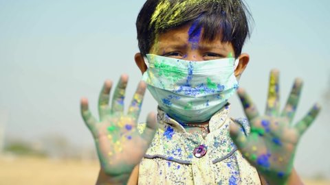 young cute little girl kid with medical face mask dancing with applied holi powder by showing to camera - concept of holi celebration during coronavirus or covid-19 pandemic with safety measures