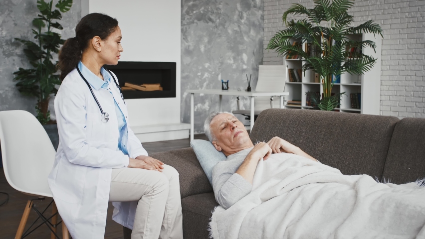 Elderly man is lying on couch and measuring temperature, giving thermometer to doctor female in uniform who saying that he has got a fever. Medical consultation at home, health check up. Close up | Shutterstock HD Video #1066404082