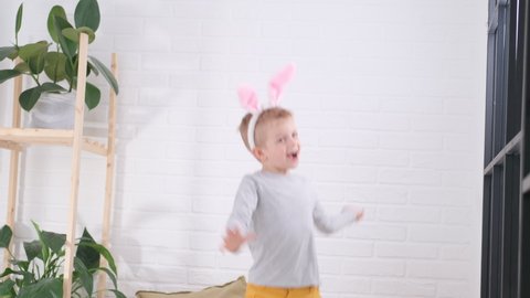 Boy in rabbit bunny ears on head jumping on white bed. Excited caucasian child in yellow pants having fun at home enjoy laughing playing funny active game in bedroom. Happy Easter celebration.