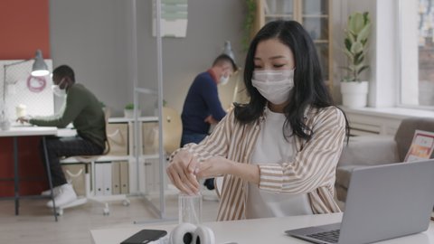 Chest up of young Asian woman with medical mask using sanitizer from bottle, then typing on laptop, sitting by desk in office. Blurred male colleagues working on background