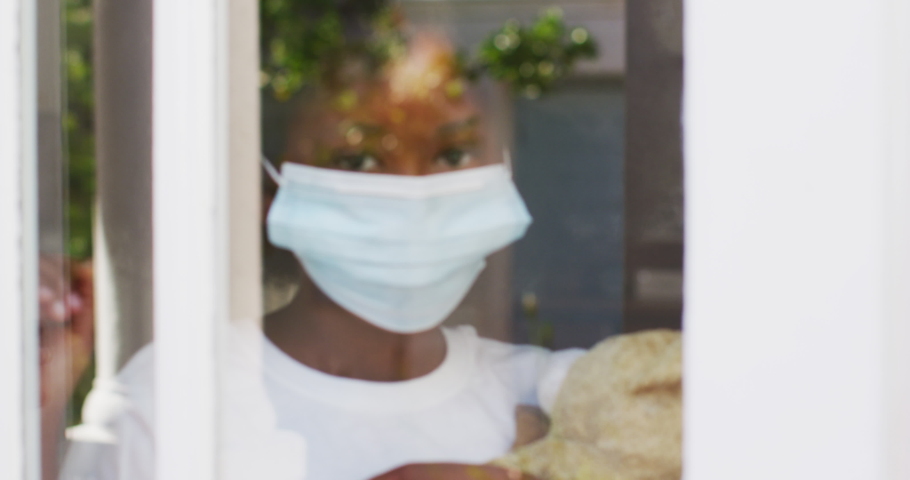 African american girl wearing face mask holding teddy bear looking through window. health and hygiene staying at home in self isolation during quarantine lockdown.