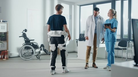 Hospital Physical Therapy: Doctor and Rehabilitation Specialist Use Tablet Computer, Helps Disabled Patient with Injury Walks while Wearing Advanced Robotic Exoskeleton Legs. Advanced Physiotherapy