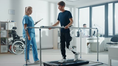 Modern Hospital Physical Therapy: Doctor Uses Tablet Computer, Helps Disabled Patient with Injury Walk on Treadmill Wearing Advanced Robotic Exoskeleton Legs. Physiotherapy Rehabilitation Technology