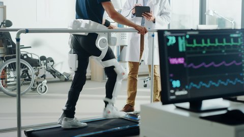 Modern Hospital Physical Therapy: Doctor Uses Tablet Computer, Disabled Patient Walks on Treadmill Wearing Advanced Robotic Exoskeleton. Computer Monitor Shows Physiotherapy Rehabilitation Activity