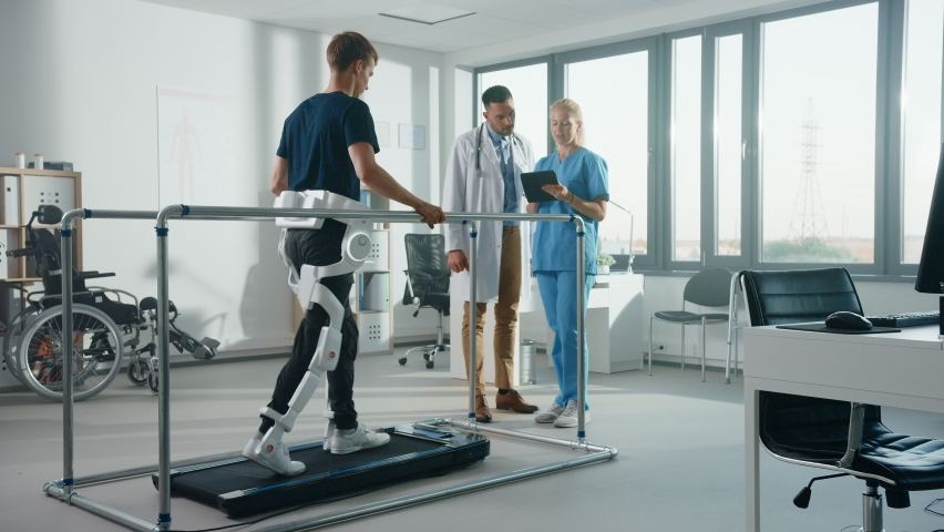 Modern Hospital Physical Therapy: Patient with Injury Walks on Treadmill Wearing Advanced Robotic Exoskeleton. Physiotherapy Rehabilitation Scientists, Engineers, Doctors use Tablet Computer to Help Royalty-Free Stock Footage #1066412728