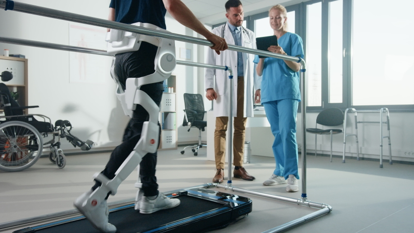 Modern Hospital Physical Therapy: Patient with Injury Walks on Treadmill Wearing Advanced Robotic Exoskeleton Legs. Physiotherapy Rehabilitation Scientists, Engineers, Doctors use Tablet Computer Royalty-Free Stock Footage #1066412731