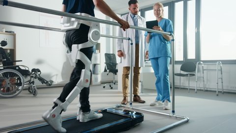 Modern Hospital Physical Therapy: Patient with Injury Walks on Treadmill Wearing Advanced Robotic Exoskeleton Legs. Physiotherapy Rehabilitation Scientists, Engineers, Doctors use Tablet Computer