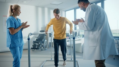 Hospital Physical Therapy: Determined Male Patient with Injury Successfully Walks Holding Parallel Bars. Physiotherapist, Rehabilitation Doctor Encourage, Applaud Disabled Person. Slow Motion