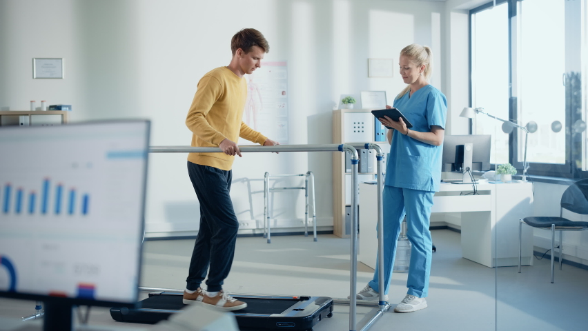 Hospital Physical Therapy Room: Patient with Injury Walking on Treadmill Holding for Parallel Bars, Professional Physiotherapist Assists, Helps, Trains Disabled Person Do Rehabilitative Physiotherapy