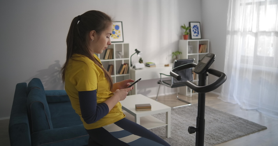 teen girl is reading messages and news in social media by cell sitting on stationary bicycle in home, sport workout for helath