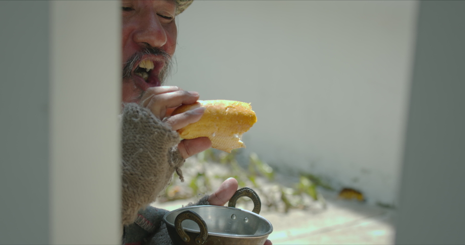 Homeless senior man with dirty clothes and old blanket wearing medical protection mask getting food from kind people and he is eating bread with happiness at the sidewalk