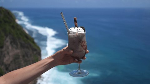 Vegan chocolate milkshake with whipped cream decorated with Oreo cookie held by hand on a beautiful ocean background