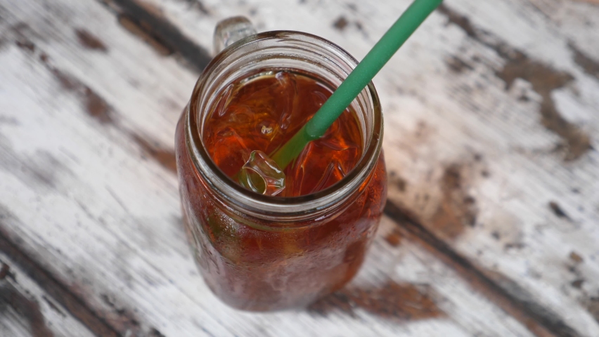 Top view of female hands stirring an ice tea in a jar with a plastic straw. Wooden table on a background  | Shutterstock HD Video #1066416451