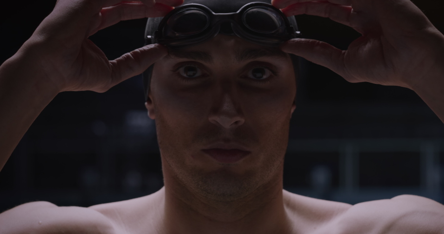 Cinematic shot of young professional male swimmer with swim cap is putting on goggles before to start practicing and training in swimming pool. Concept of sport, competition, victory, fitness, passion | Shutterstock HD Video #1066418488