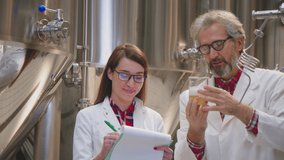Two food technologists in white lab coats discussing and looking at a beer sample and taking notes. Man holding beer sample in his hands, woman smiling and taking notes. Science, brewery concept.