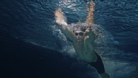 Cinematic shot of young professional male swimmer with cap and goggles trains with effort and dedication to win the race by practicing in swimming pool. Concept of sport, competition, victory, fitness