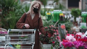 Blonde woman in face mask and coat is chooses flowers in a garden store 