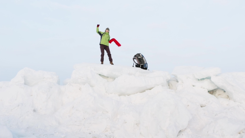 Zooming in view from below of a happy woman dancing and waving the red scarf on big icy blocks against the blue sky. Winter