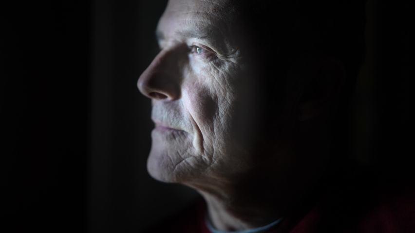 Dramatic portrait of an attractive mature man in a low light who is looking out the window and then looking at the camera. Black background. Royalty-Free Stock Footage #1066455220