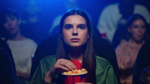 Portrait of young female person watching movie in dark hall. Attractive girl with long hair eating popcorn in movie theater in slow motion. Closeup beautiful woman enjoying interesting film in cinema.
