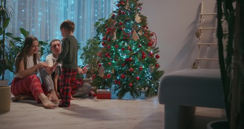 A family with two children sit together at the Christmas tree and cuddle. Love and family happiness on Christmas Eve. Father and two sons cuddle and look at Christmas tree in their living room