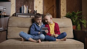 Pleasant satisfied small two boys in casual clothes resting on soft couch in contemporary room and using tablet device to watch cartoon or play video game