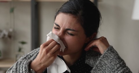 Close up head shot unhealthy millennial indian mixed race woman covered in warm plaid sneezing in paper tissue, having infectious disease, caught cold or suffering from grippe symptoms at home.