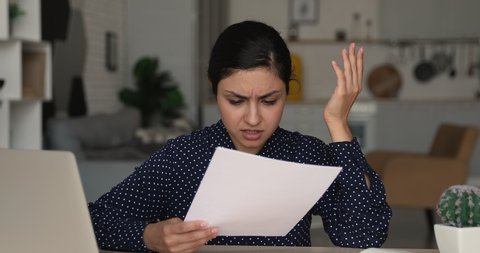 Focused young 25s indian ethnicity woman holding paper document in hands, feeling unhappy with exam failure, received negative news notification in correspondence, eviction notice or dismissal letter.