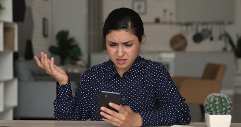 Stressed young multiracial indian woman typing using cellphone applications, feeling confused of bad electronic device work or getting message with unpleasant news, sitting at table at home office.