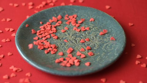 Red scarlet heart shaped sugar confetti crumbling and falling down on green plate on red tablecloth cloth. Romantic love, affection or Saint Valentine's Day preparation design concept