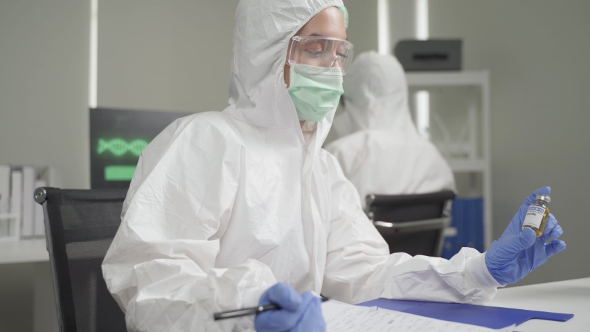 Young female scientist in PPE,mask,protective devices is sitting in modern sophisicated lab and holding up a corona virus vaccine bottle.She gives the medicine to a male scientist and make discussion. | Shutterstock HD Video #1066477114