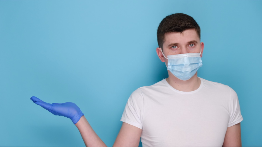 Young male holds paper white arrow, wears respirator face mask and latex gloves to protect herself from coronavirus, demonstrates empty copy space for promo text, model over blue background studio