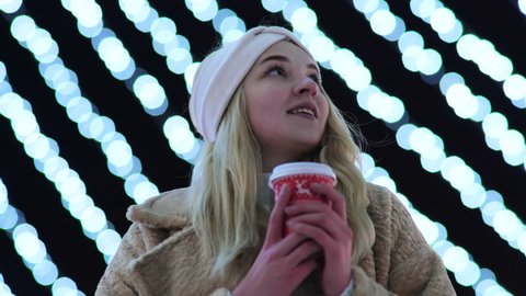 A girl with a cup of warm drink on the background of the Christmas lights of the evening city.