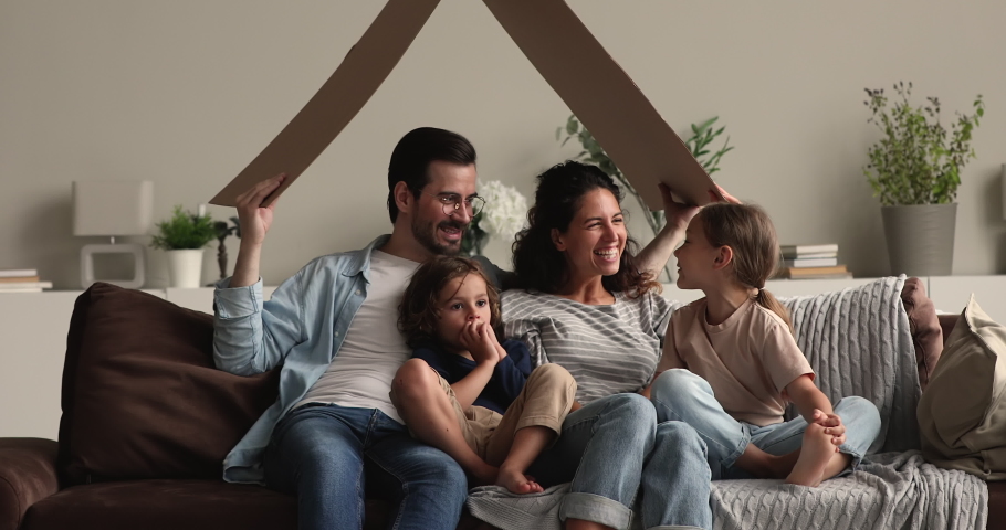 Beautiful young married couple with little son and daughter sitting together on couch under carton roof, symbol of tenancy, bank loan, happy homeowners family, new real estate buyers portrait concept Royalty-Free Stock Footage #1066486753
