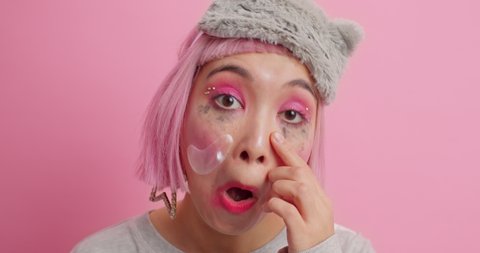 Asian woman with bob hairstyle looks attentively at her face examines skin wipes leaked makeup under eyes prepares for sleep wears soft blindfold poses against pink background. Beauty concept
