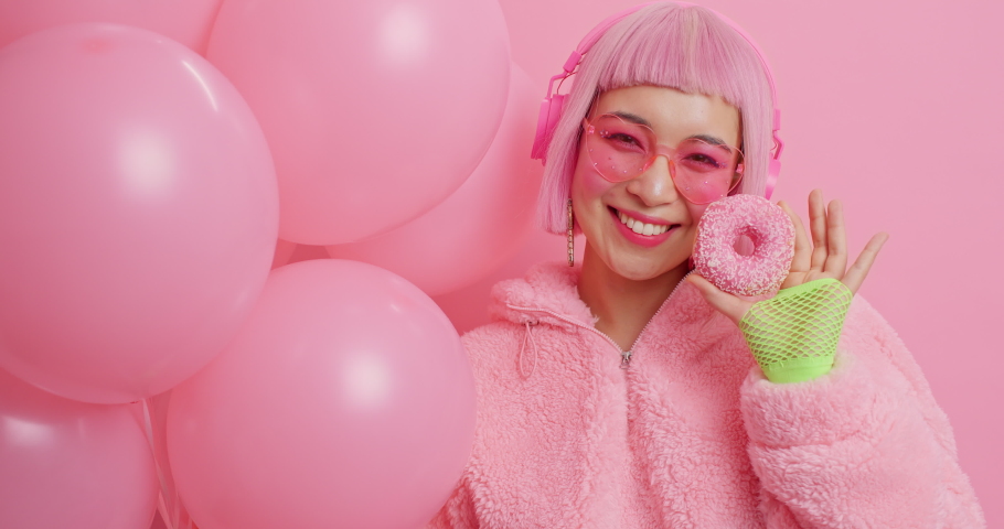 Pleased young Asian woman with pink hair dressed in fur coat holds delicious doughnut and bunch of inflated of balloons isolated over pink background celebrates special occasion in her life.