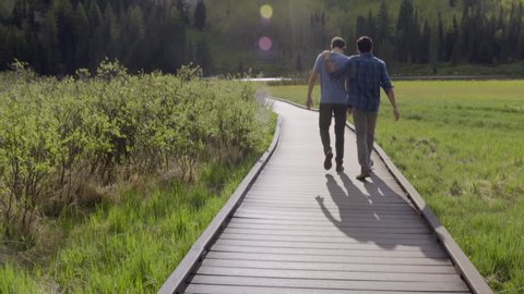 Best Friends Walk Down Boardwalk Toward Mountain Lake, Boy Puts His Hand On His Friend's Back 
