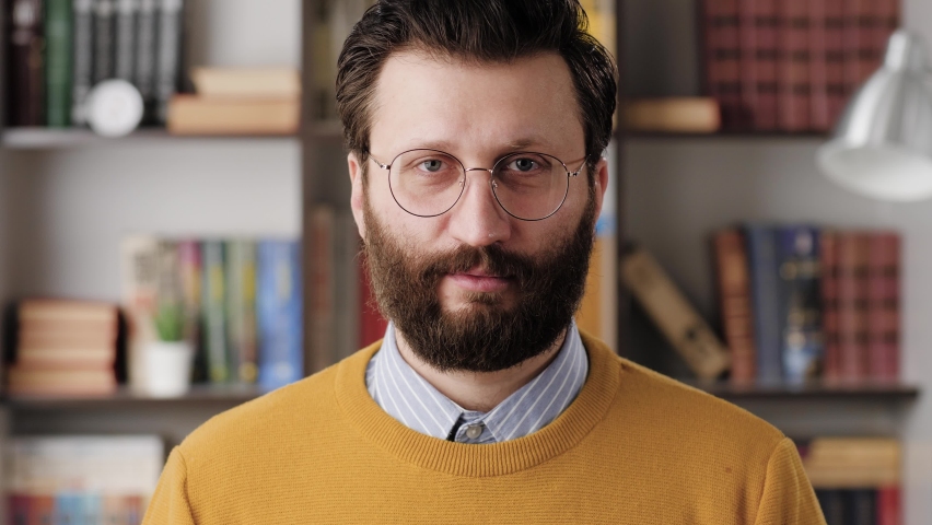 Man is surprised, shocked. Confused bearded man in glasses in office or apartment room looking at camera and slowly takes off his glasses in surprise looking at camera. Close up and slow motion