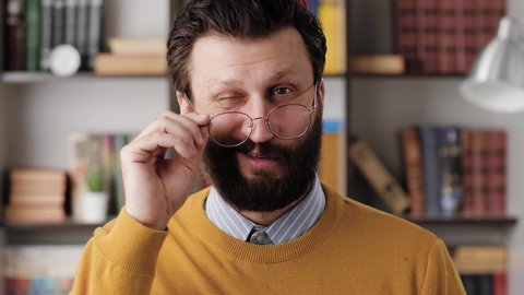 Man winks. Positive smiling bearded man with glasses in office or apartment room looking at camera and slowly lowers his glasses and winks. Close up and slow motion