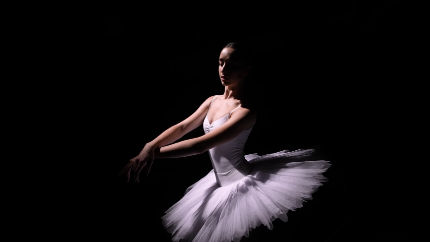 Camera rotates around gentle ballerina in a white tutu performing graceful movements with her hands. Orbital shot of a young dancer soaring against a black studio background. Close up. Slow motion.