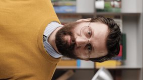 Man is surprised, shocked. Vertical video of confused bearded man in glasses in office or apartment room looking at camera and slowly takes off his glasses in surprise looking at camera. Close up