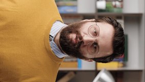 Man is praying. Vertical video of upset bearded man with glasses in office or apartment room looking at camera and brings his hands to his face and begins to say prayer. Close-up and slow motion