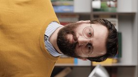 Man beckons with his finger. Vertical video of serious frowning bearded man with glasses in office or apartment room looking at camera and points his fingers gesturing to him. Close-up and slow motion