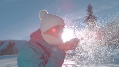 SUPER SLOW MOTION, CLOSE UP, LENS FLARE, PORTRAIT, DOF: Carefree young woman blows into her hand full of snow and turns to the camera. Playful female tourist blows sparkling snowflakes in the air.