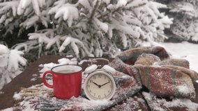 Vintage alarm clock and cup of coffee on wooden table in snowfall near pine tree